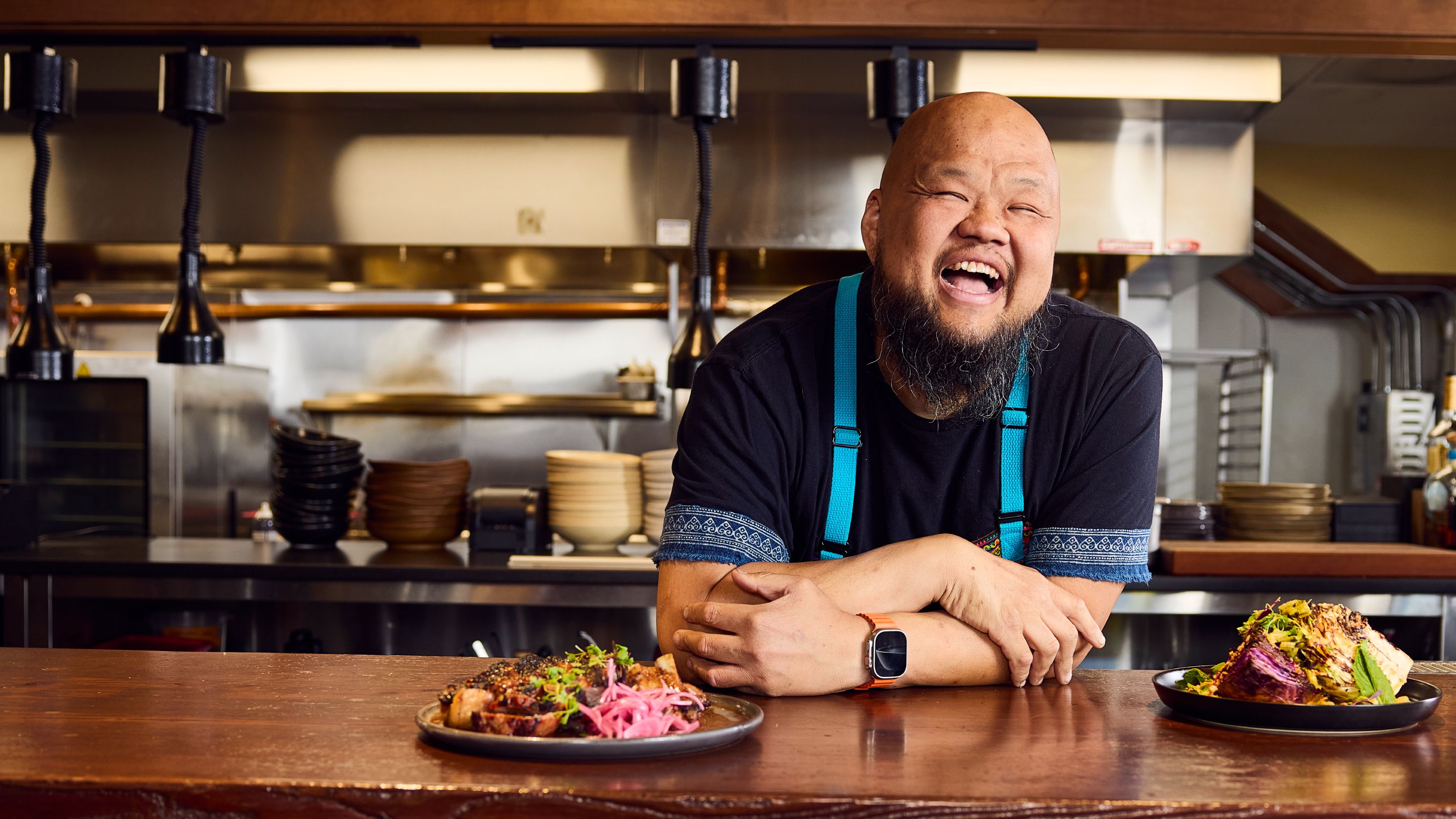 Photo of Chef Yia Vang leaning on a counter with a plate of food on it and in the background is the kitchen at his restaurant Vinai. 